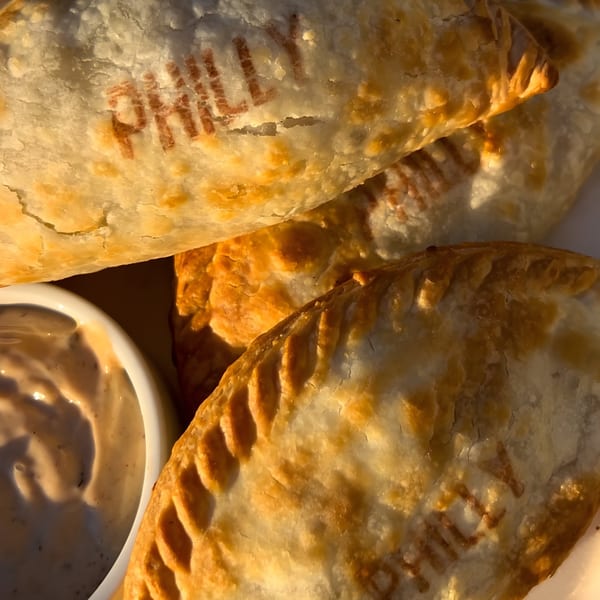 Close-up of golden-brown Nosh Empanadas, one stamped "PHILLY," with a side of dipping sauce.