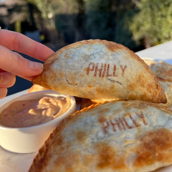 Hand holding a "Philly" empanada above a dipping sauce, with more empanadas visible.