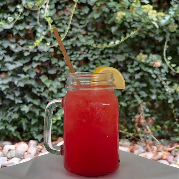 A vibrant red drink in a mason jar with a lemon slice and straw, set against a leafy green background.