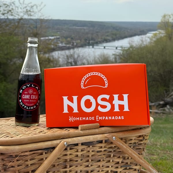 Nosh Empanadas box and Boylan Cane Cola on a picnic basket with a river view.