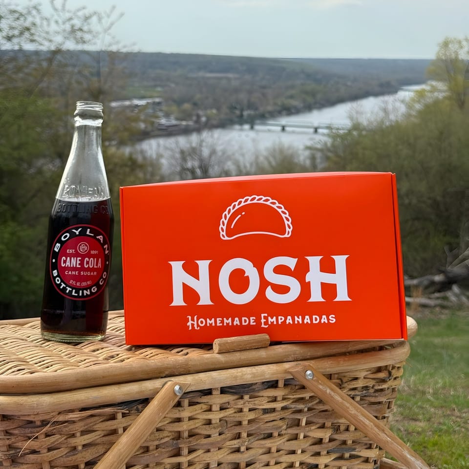 Nosh Empanadas box and Boylan Cane Cola on a picnic basket with a river view.